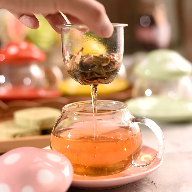 Person pouring tea from a glass teapot into another glass cup on a blurred background.