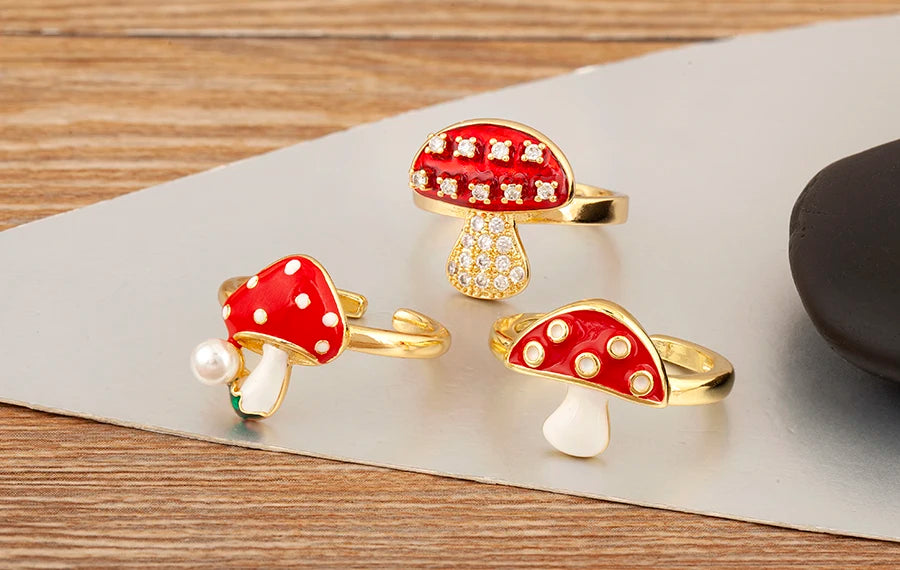 Three mushroom-shaped rings with red caps and gold bases on a wooden surface.