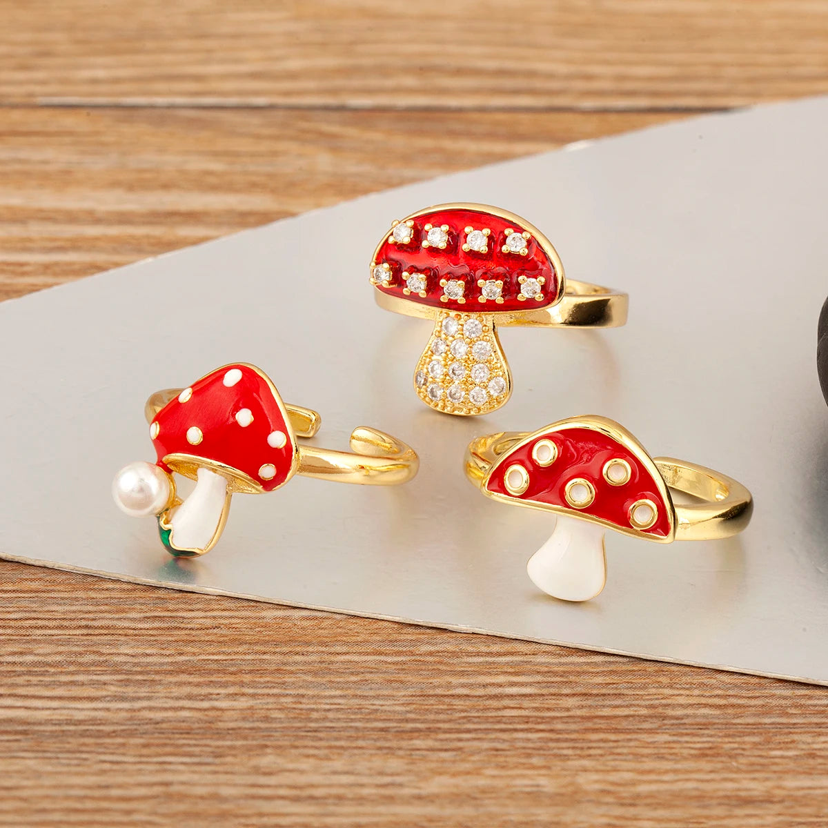 Three mushroom-shaped rings with red caps, white spots, and gold bases on a wooden surface.