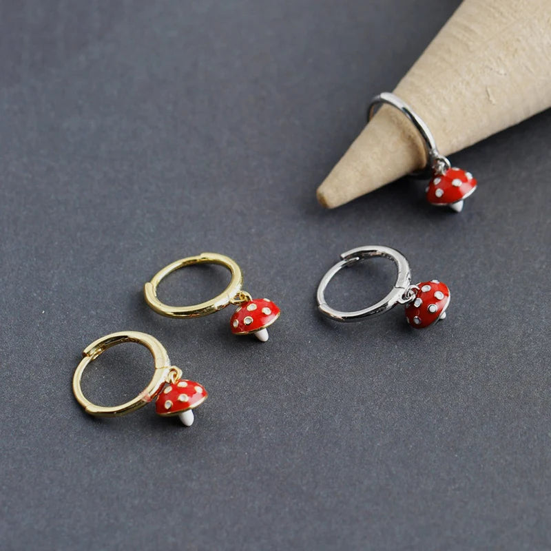 Two pairs of hoop earrings with red mushroom charms on a gray surface.