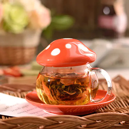 Glass teapot with red mushroom lid on a saucer, placed on a woven surface.