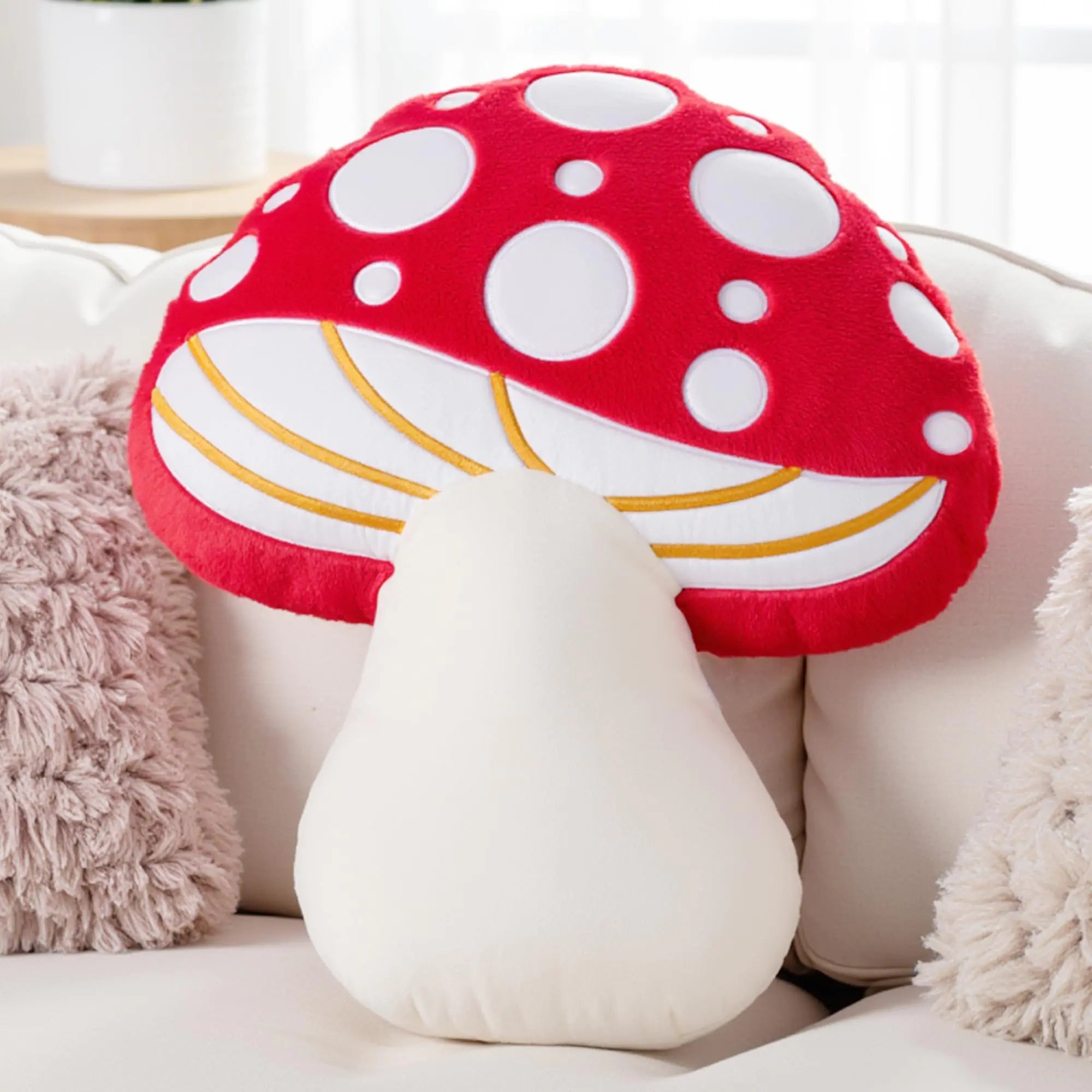 Mushroom-shaped pillow with red cap and white spots on a light-colored couch.