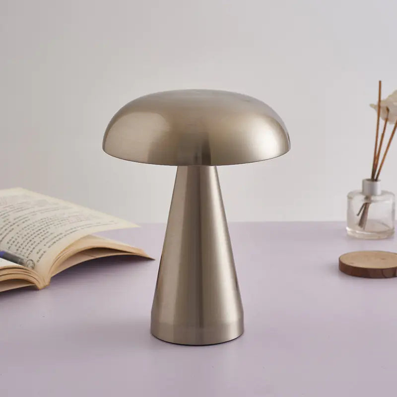 Gold mushroom-shaped table lamp on a light purple surface with an open book and small plant in the background.